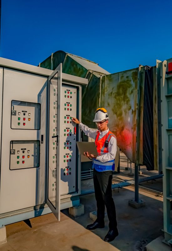 A worker in a safety helmet and vest inspects an electrical control panel outside a utility facility.